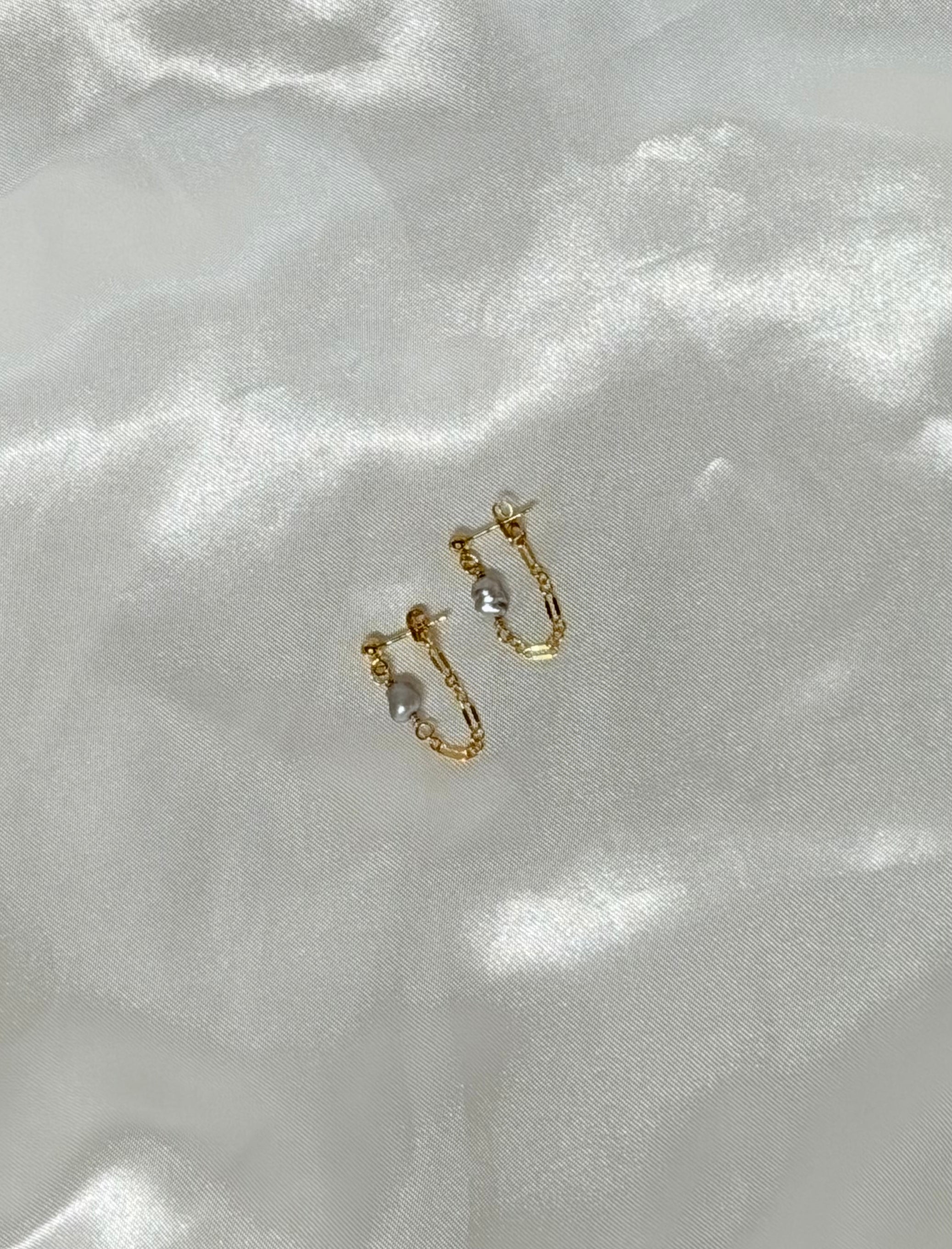 A pair of 14k gold-filled chain loop earrings with a gray keshi pearl, displayed on white satin fabric in natural light.