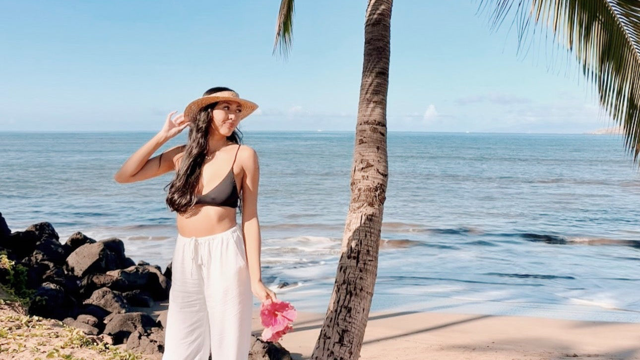 Woman in a bikini and hat standing on a beach with palm trees and ocean in the background