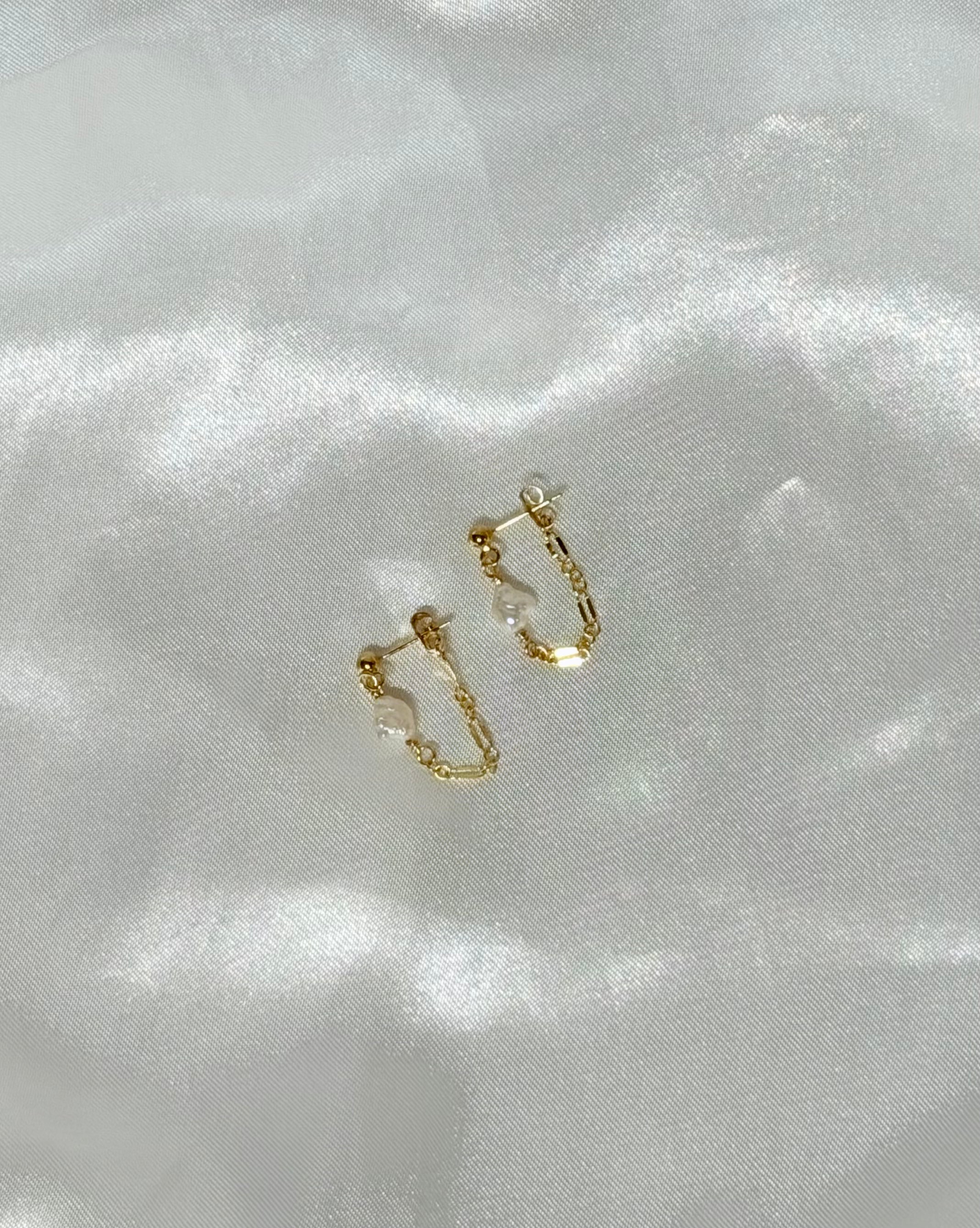 A pair of 14k gold-filled chain loop earrings with a white keshi pearl, displayed on white satin fabric in natural light.